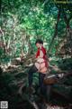 A woman in a red shirt and black stockings posing in the woods.