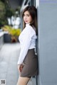 A woman leaning against a wall in a white shirt and brown skirt.